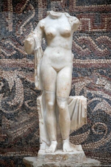 Headless Roman marble female nude with side drapery and support, displayed before a patterned mosaic.