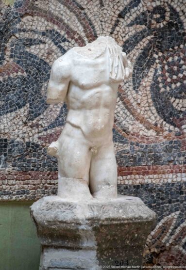 Fragmentary Roman marble male nude torso missing the head and arms, mounted against a patterned mosaic wall.