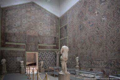 Djemila Museum corner with monumental geometric mosaics on two walls, a central marble statue, smaller sculptures, and a case.
