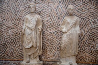 Two draped Roman marble statues standing side by side in front of a multicolored geometric mosaic.