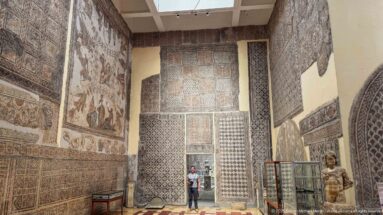 Visitor standing in a tall doorway surrounded by figurative and geometric Roman mosaics in Djemila Museum.