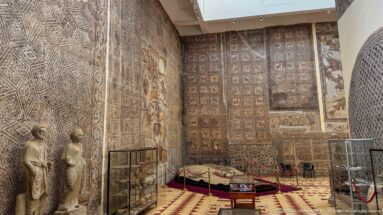 Djemila Museum gallery with two draped Roman statues, large mosaic-covered walls, display cases, and a roped platform.