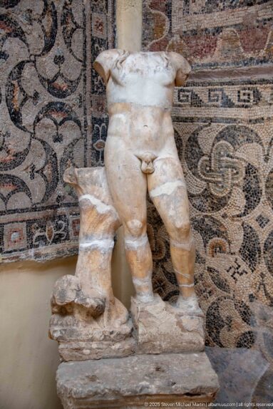 Fragmentary marble nude male statue missing its head and arms, displayed in front of a geometric Roman mosaic.