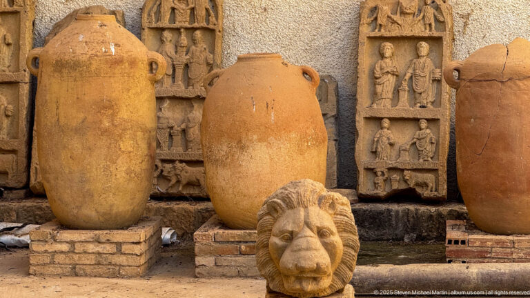 Large Roman amphorae standing before carved relief plaques, with a sculpted lion head in the foreground.