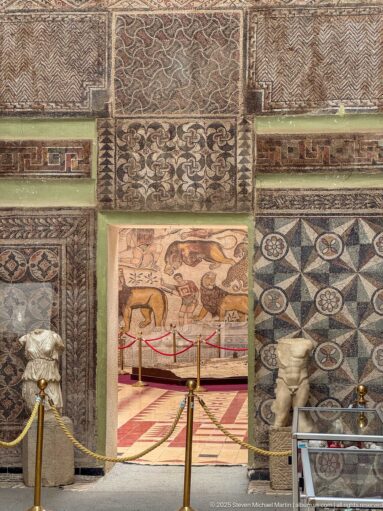 Mosaic-framed doorway between Djemila Museum galleries, with geometric panels, floral borders, and sculptures at floor level.