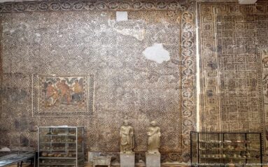 Djemila Museum gallery with large wall-mounted Roman mosaics, two statues, and display cases of archaeological objects.