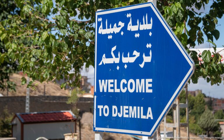 Blue roadside sign reading Welcome to Djemila in Arabic and English.