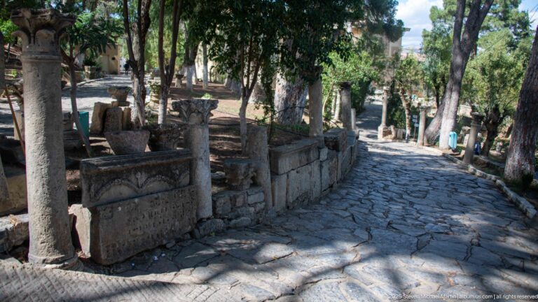Stone-paved path under trees with ancient blocks and column fragments displayed along both sides.