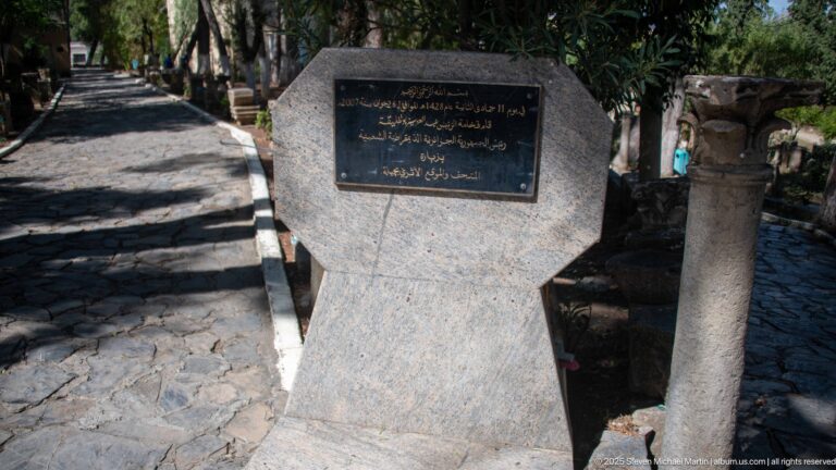 Dark Arabic inscription plaque on a polished stone pedestal beside a weathered column.