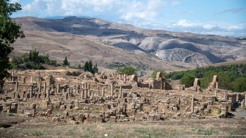 Broad panorama of Djemila's stone ruins and columns with layered mountain ridges beyond.