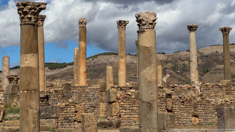 Group of tall Roman columns, some with carved capitals, rising above low masonry walls at Djemila.