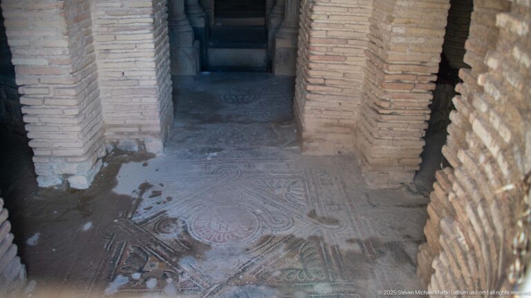Geometric mosaic floor with circular motif inside a narrow Roman brick interior with arched openings.
