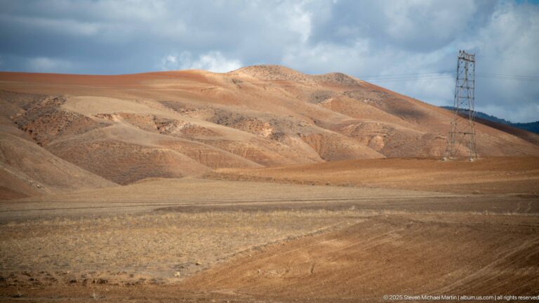 Dry ochre hills with layered slopes and a tall electrical transmission tower near Djemila.