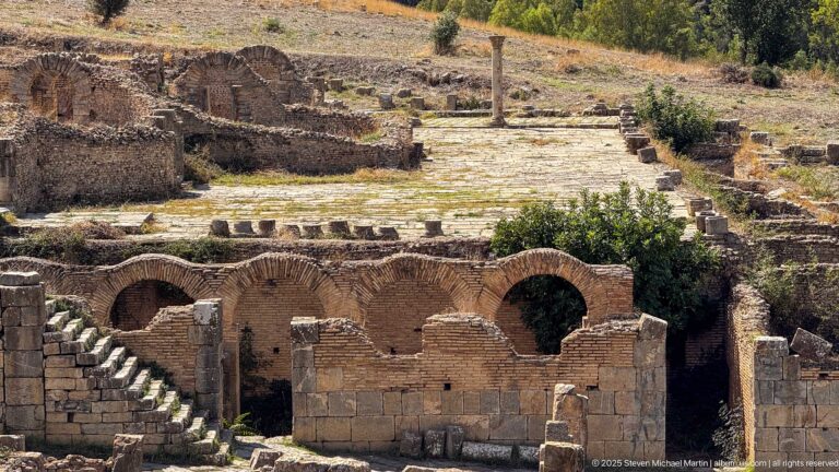 Low Roman brick façade with repeated arched openings beside a stone-paved terrace and scattered ruins.