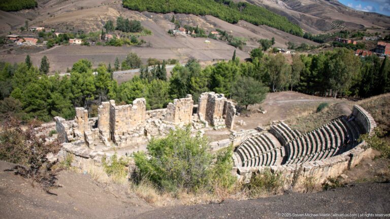 Elevated view of Djemila's Roman theater with semicircular stone seating, orchestra, and ruined stage building.