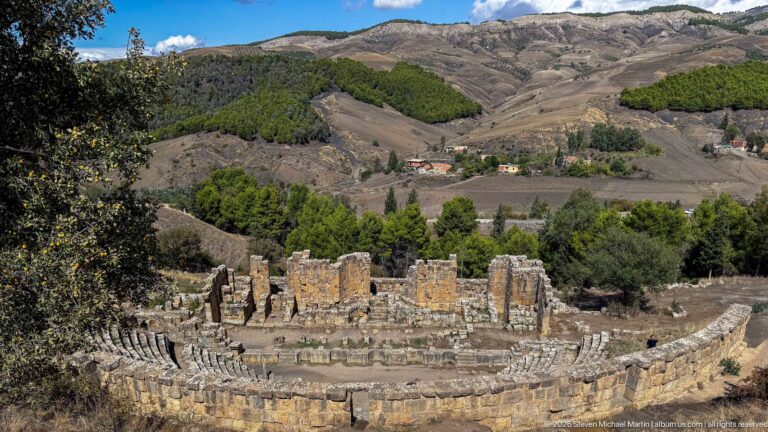 Wide view across the curved seating and stage remains of Djemila's Roman theater against steep hills.
