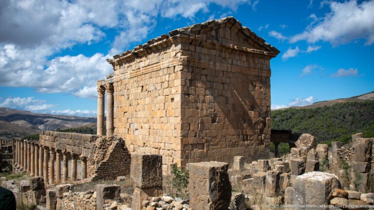 Oblique view of a large Roman podium temple with pediment and side colonnade among Djemila's ruins.