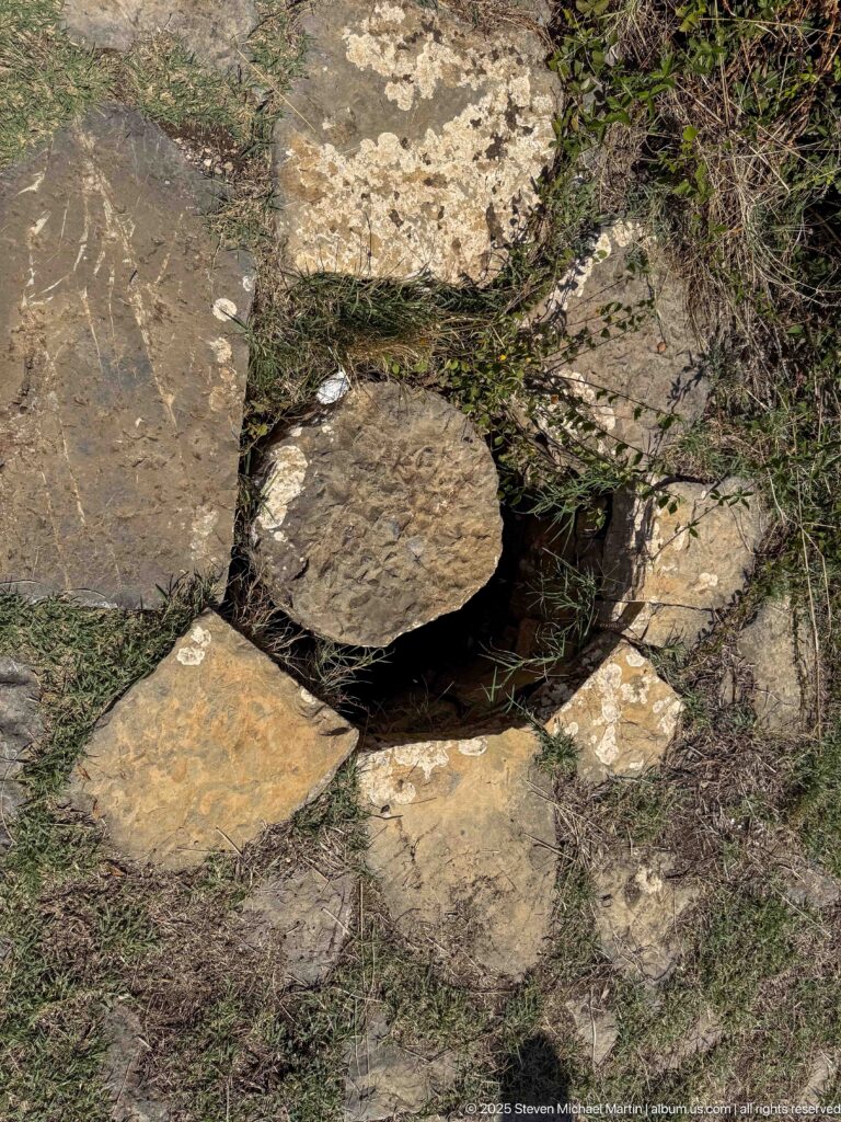 Round stone disk set among large weathered paving slabs with grass growing in the joints.