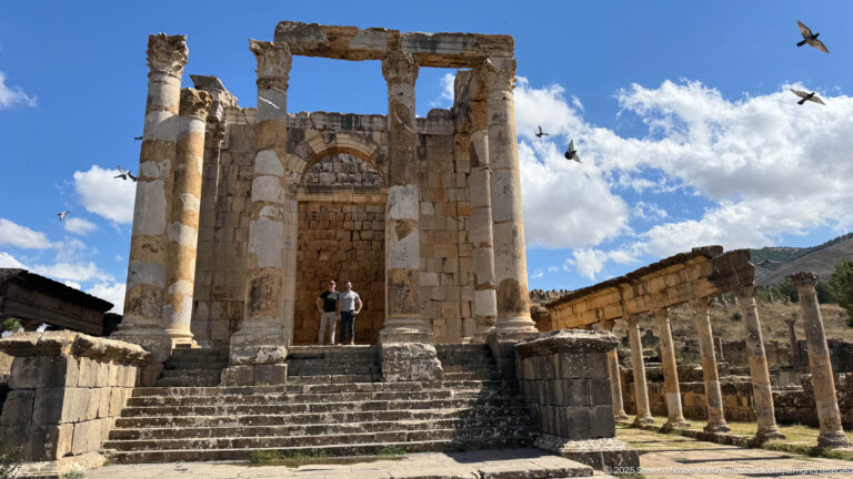 Two visitors standing on the steps of a large Roman temple with columns and adjoining colonnade at Djemila.