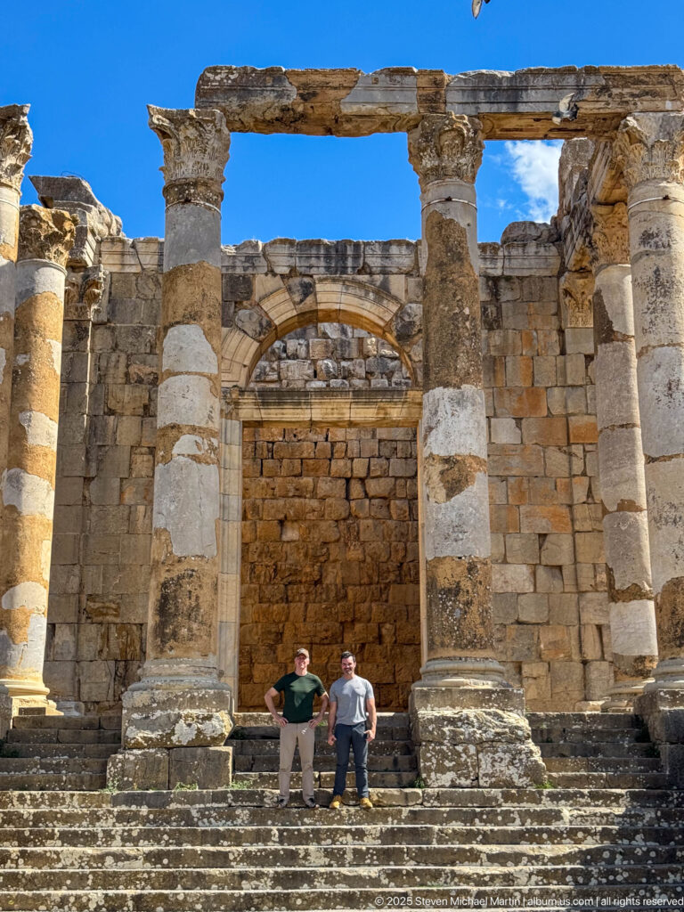 Two visitors on the broad steps beneath massive columns and masonry of a Roman temple at Djemila.