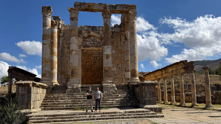 Two visitors at the foot of a monumental Roman temple stair with columns and long side colonnade.
