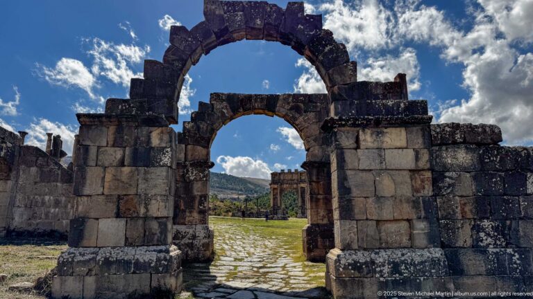 Large and smaller aligned stone arches framing a paved route and distant Roman monument at Djemila.