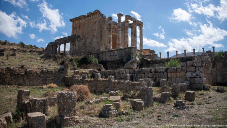 Roman temple rising above terraced ruins, scattered blocks, and column fragments at Djemila.