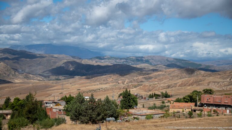 Wide Djemila highland valley with scattered buildings, trees, fields, and layered hills.
