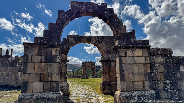 Large stone arch framing a smaller opening and a distant Roman triumphal arch at Djemila.