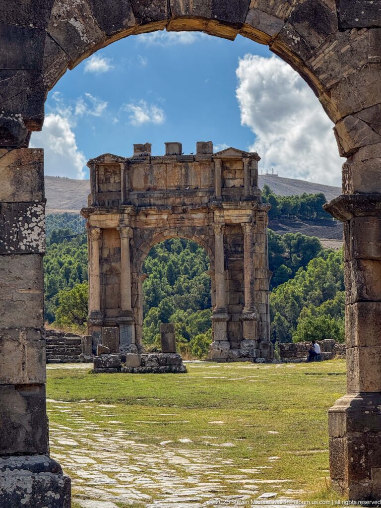 Roman triumphal arch centered through a foreground stone arch, with green hills beyond at Djemila.