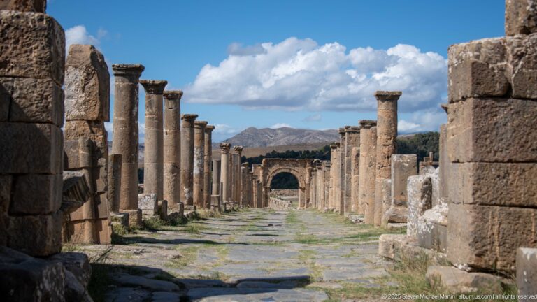 Long stone-paved Roman street lined with columns and walls, terminating in a distant arch at Djemila.