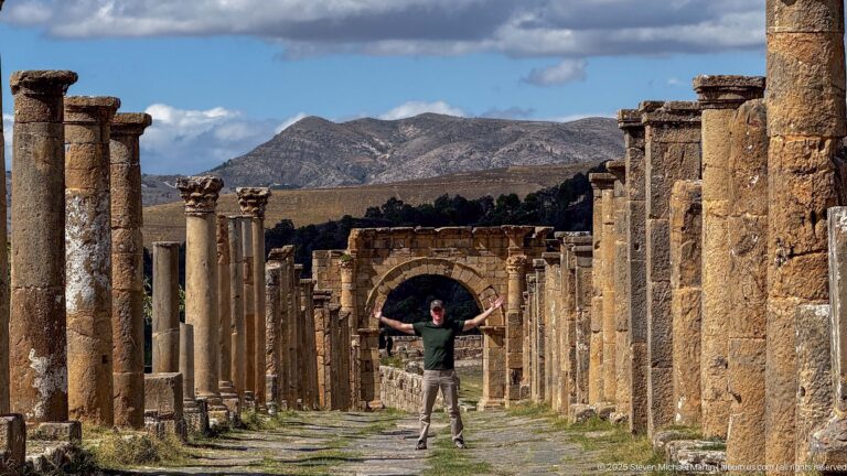 Visitor with outstretched arms standing on a long Roman street lined with columns and walls at Djemila.