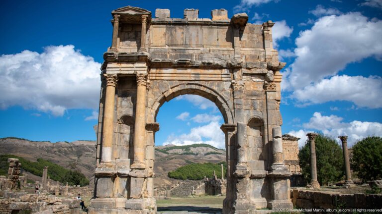 Monumental Roman triumphal arch with broad central opening, paired columns, and mountain backdrop at Djemila.