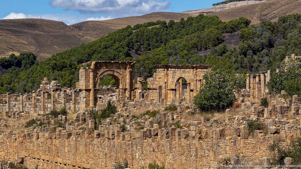 Large Roman arched structure rising among dense columns, walls, and masonry ruins at Djemila.