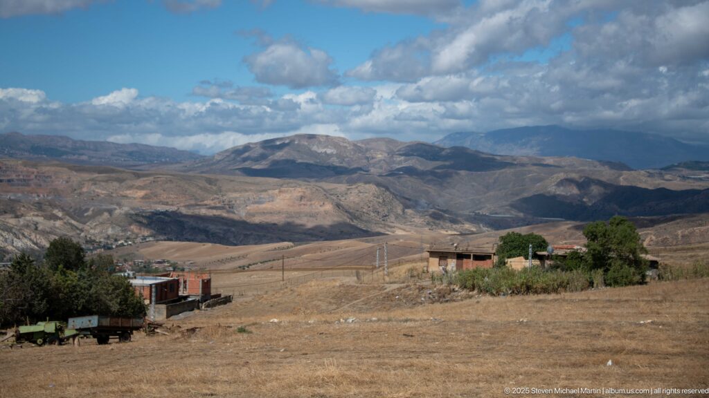 Rural outskirts of Djemila with low buildings, trees, open ground, and layered mountains.