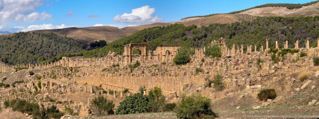 Wide hillside panorama of Djemila with dense Roman columns, walls, monuments, and wooded slopes.