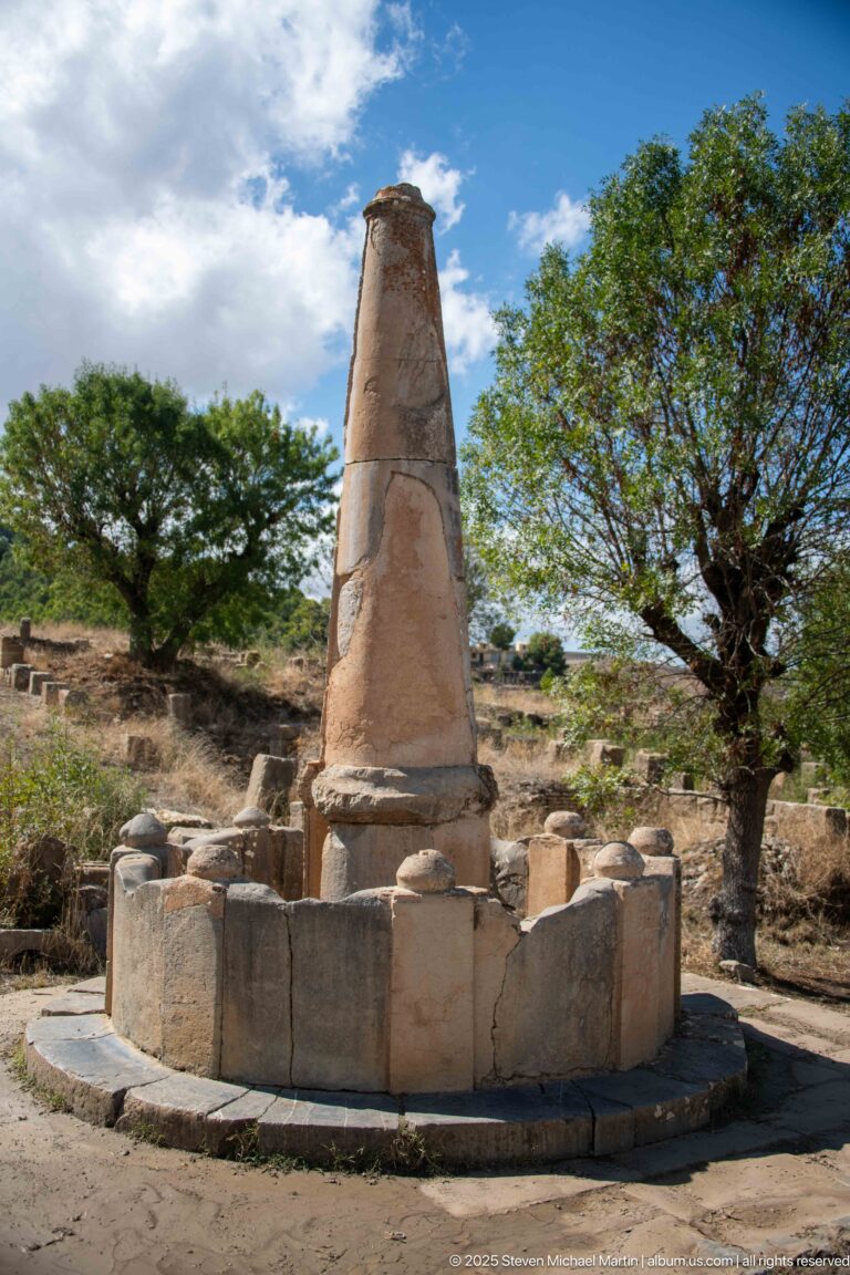 Tall tapered stone monument standing on a low circular block enclosure among trees at Djemila.