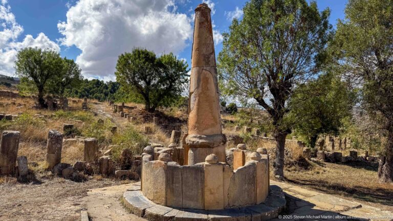 Wider view of a tall tapered stone marker in a circular enclosure amid trees and scattered ruins at Djemila.