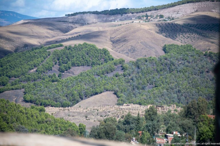 Distant Djemila ruins appearing as a pale stone complex within green and cultivated hills.