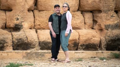Two visitors standing together before massive weathered dressed-stone blocks in the exterior wall of the Medracen.