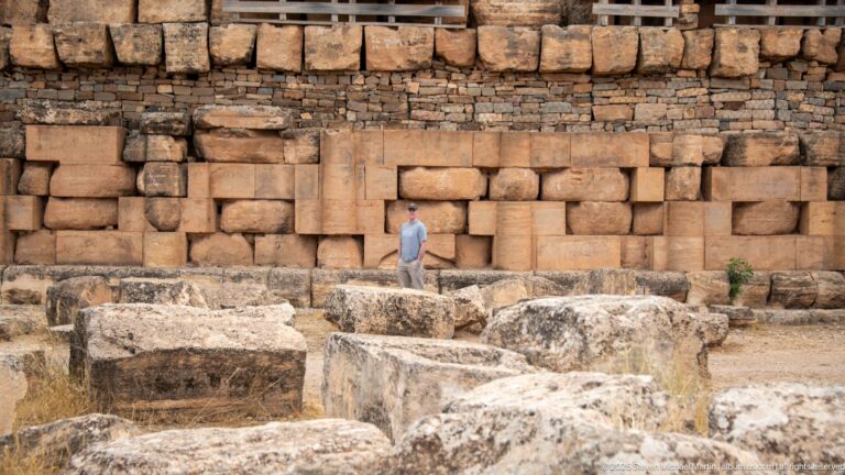 Visitor standing before the Medracen’s engaged-column wall, with massive displaced stone blocks scattered across the foreground.