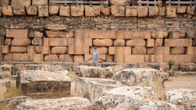 Visitor standing before the Medracen’s engaged-column wall, with massive displaced stone blocks scattered across the foreground.