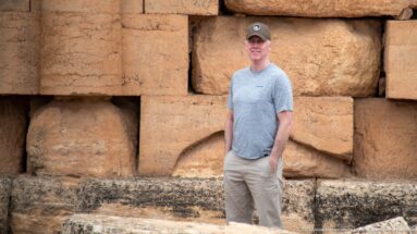 Man in gray shirt and cap standing beside massive weathered stone blocks in the exterior wall of the Medracen.