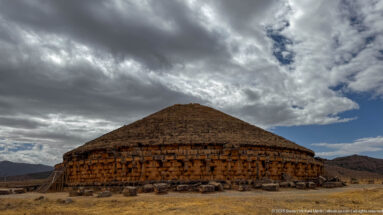 Medracen mausoleum under dramatic dark clouds, with warm light on the circular stone wall and distant hills at right.