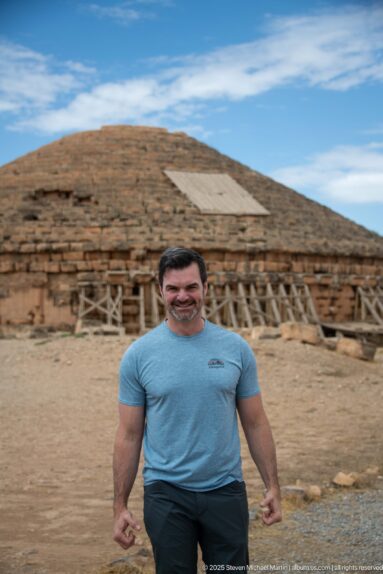 Man in blue shirt standing before the Medracen, with its stepped roof, circular masonry wall, and timber braces behind him.