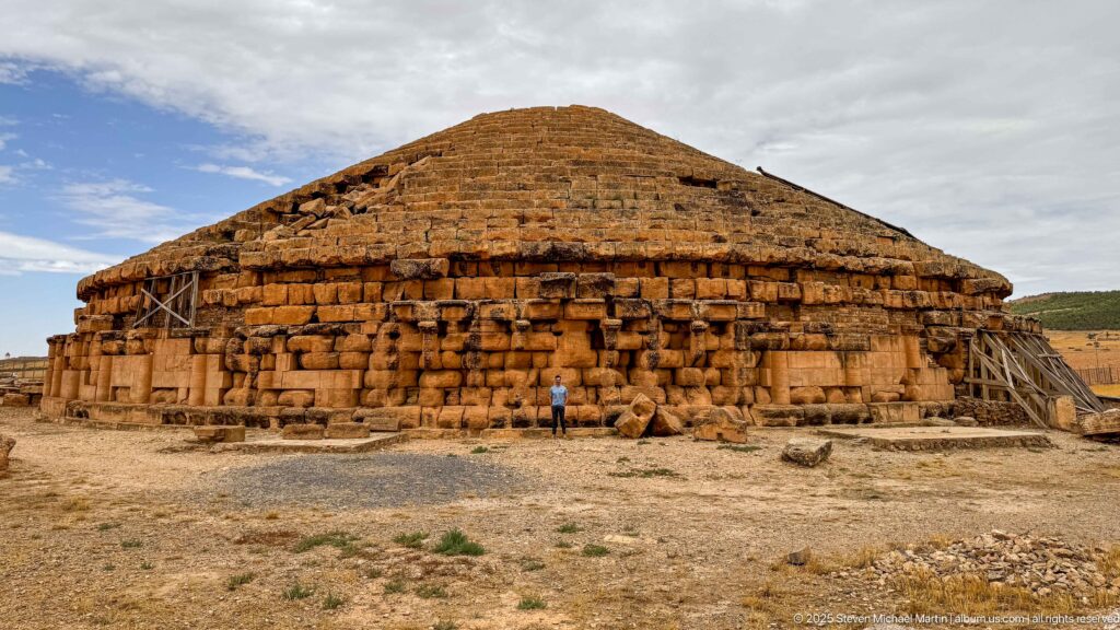 Visitor standing before the massive circular Medracen mausoleum, whose stepped roof and damaged stonework are supported by timber braces.