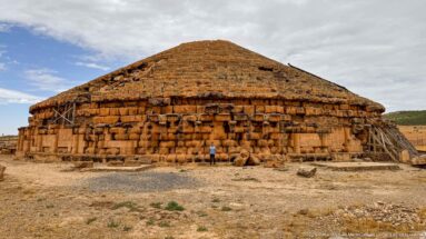 Visitor standing before the massive circular Medracen mausoleum, whose stepped roof and damaged stonework are supported by timber braces.