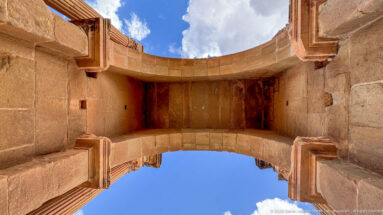 Upward view beneath the Arch of Trajan at Timgad, showing the stone vault, piers, cornices, and blue sky.