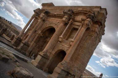 Oblique low-angle view of Timgad’s Arch of Trajan with central arch, side opening, columns, and niches.