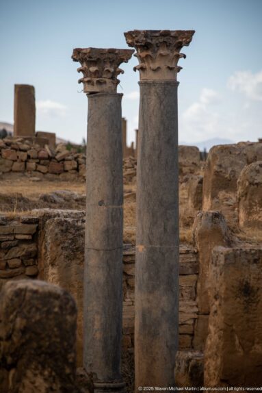 Two standing stone columns with carved capitals rising above low archaeological ruins at Timgad.